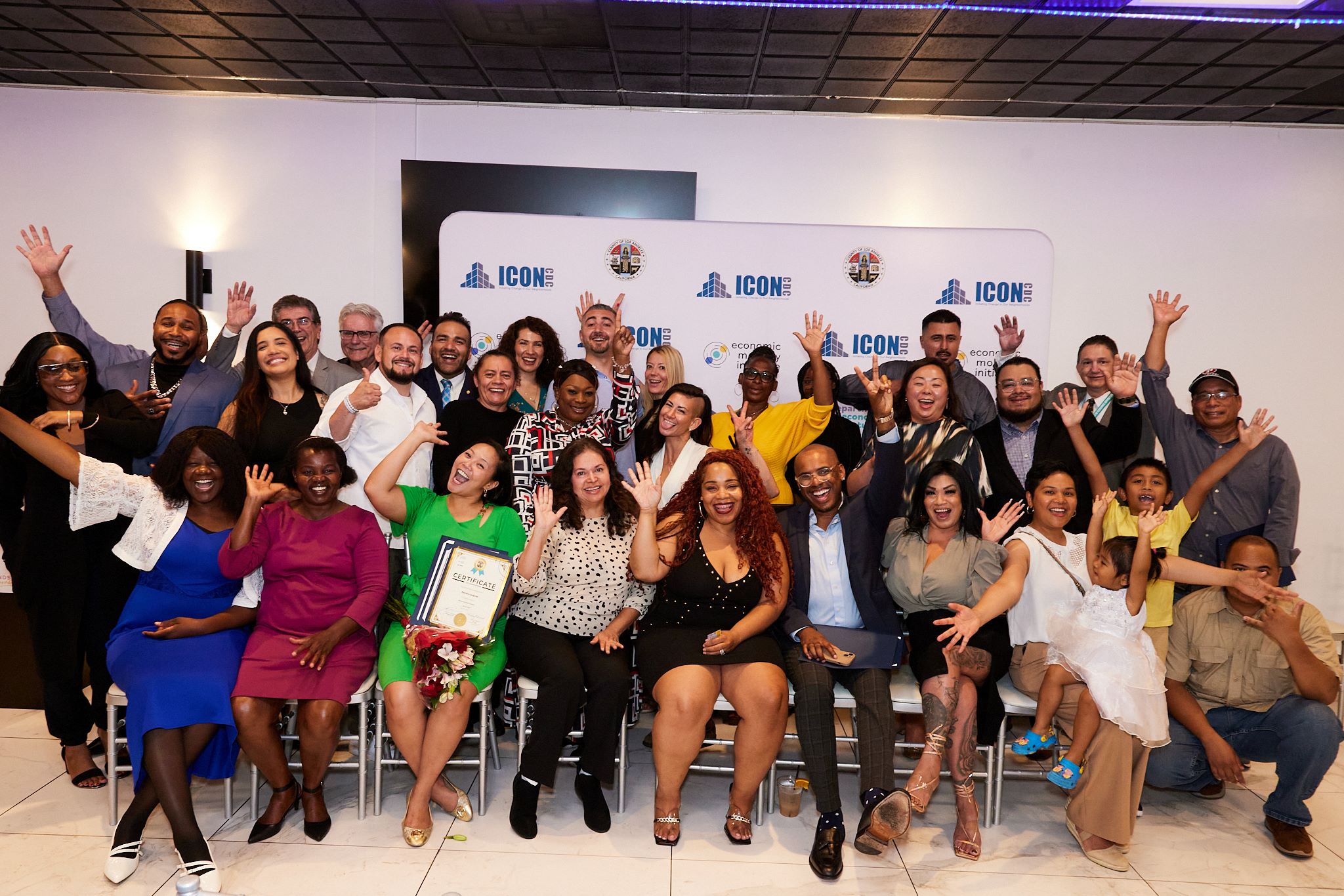 A large group of people is gathered together celebrating at what appears to be a graduation or recognition event, with many smiling, raising their hands, and posing enthusiastically in front of a backdrop displaying the logos of ICON CDC, the County of Los Angeles, and the Economic Mobility Initiative.