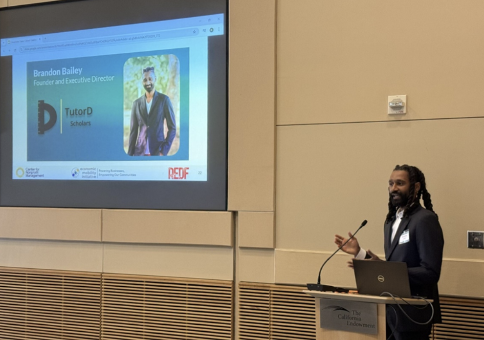 Presenter at a podium beside a projected slide for TutorD Scholars showing a headshot and the title ‘Founder and Executive Director.